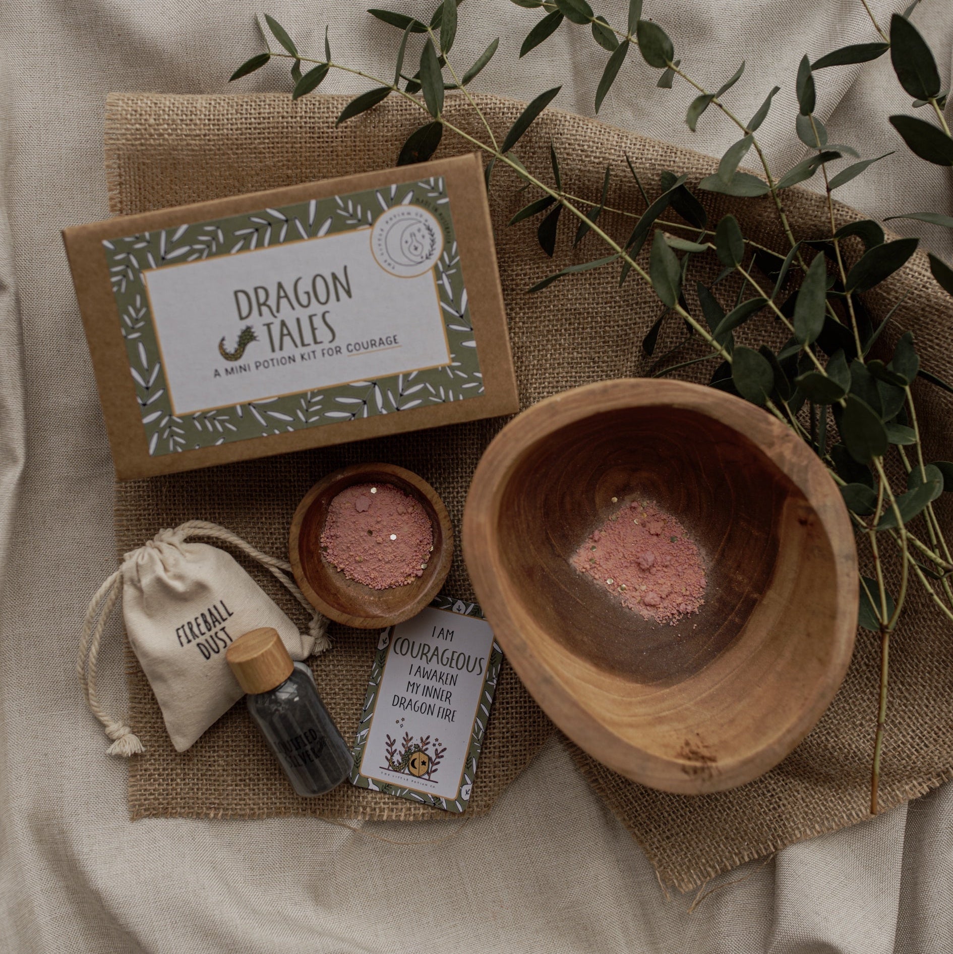 A styled flat-lay of a 'Dragon Tales' bath soak kit on a beige fabric background with burlap and eucalyptus. The kit includes a kraft paper box, a small wooden bowl filled with pink bath salts, a small fabric pouch, a small glass bottle, and a wooden spoon, all arranged around a larger wooden bowl.